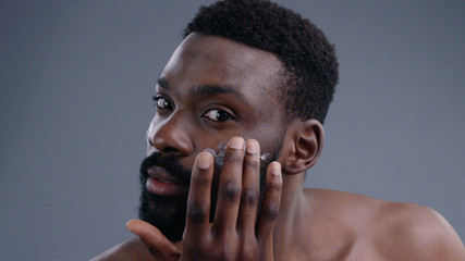 Portrait of attractive african young man using a moisturizing facial cream gently touching his face looking in the mirror. Close-up of nice-looking guy demostrating a jar of cream smiling at camera.