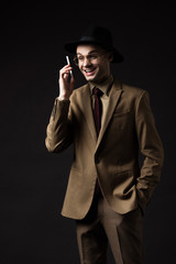 Obraz premium smiling elegant man in beige suit, hat and eyeglasses talking on smartphone isolated on black