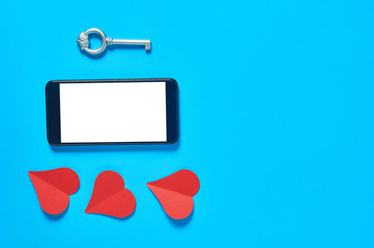 Black smartphone with isolated white screen for text, picture, photo and other graphics near paper hearts and vintage key on blue table. Secrets or forbidden love concept. Space for text. Top view