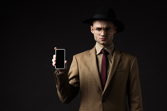 serious elegant man in beige suit, hat and eyeglasses holding smartphone with blank screen isolated on black