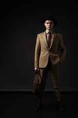 elegant man in beige suit, hat and eyeglasses holding brown leather bag with hand in pocket on black