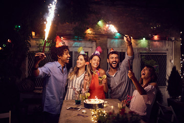 Smiling group of young friends having  birthday party in the club with cake and candles at night.  Party, fun, birthday concept. - obrazy, fototapety, plakaty