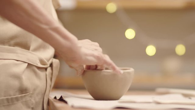 Close-up shot with bokeh of wrinkly hands of anonymous senior lady in beige apron making clay bowl, sculpting walls and smoothing outer surface with thumb