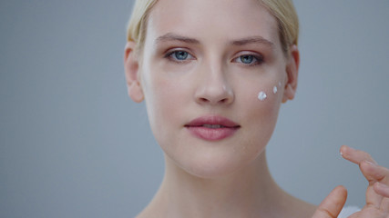 Glorious happy blonde woman applying skincare lotion on face in a bathroom in front of a mirror....