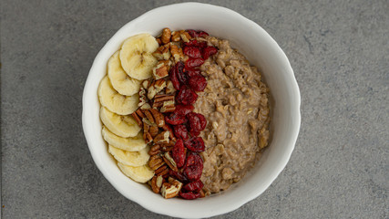 Breakfast oatmeal with banana, pecan nuts, dry cranberry in white bowl on a grey background