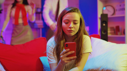Lonely pretty girl getting bored on friends party, sitting on couch browsing social media while other people having fun celebrating holiday. Concept of nomophobia.