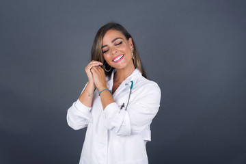 Dreamy young doctor woman with pleasant expression, closes eyes, keeps hands crossed near face, thinks about something pleasant, poses against gray background.