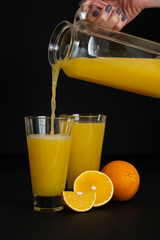 the hand of a young woman pours orange juice from a jug into a glass, black background, vertical frame, copy space