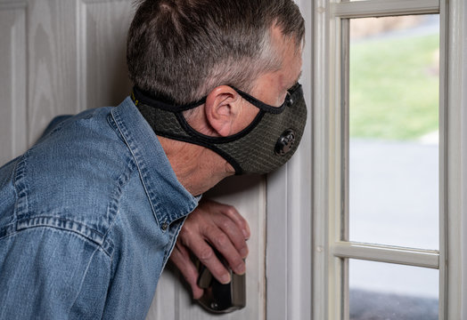 Concerned And Worried Man Wearing A Protective Breathing Mask Against Flu And Coronavirus And Looking Out Of Front Door