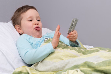 The child is lying on the bed, sick. The child refuses to be treated with pills. The child shows his tongue in disagreement.