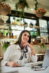 Beautiful caucasian businesswoman sitting in cafe, taking a break from hard work and holding cup of coffee. On table is laptop.