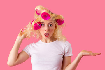 Studio portrait of an excited young blonde woman with hair curlers and open mouth, isolated on pink
