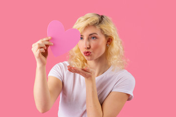 Studio portrait of a young casual woman holding pink heart and blowing kiss with lips pouted