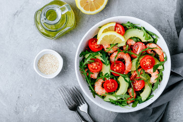 Healthy arugula salad bowl with shrimp, avocado, tomato, and sesame seeds on gray stone background.