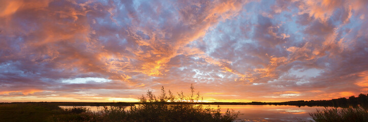 Panorama of bright colorful sunset on the background of yellow-red clouds reflected in water of lake