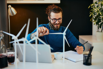Young creative caucasian bearded environmentalist  with curly hair sitting in his office, touching windmill model and drawing new better model in notebook.