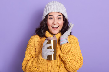 Smiling astonished woman holding thermo mug with hot beverage and spreading palm aside, posing isolated over blue studio background, lady wearing winter clothing, having surprised expression.