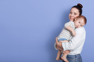 Horizontal shot of beautiful young mother wears casual clothing with bun standing and holding her baby girl in her arms isolated over blue studio background. Copy space for advertisment or promotion.