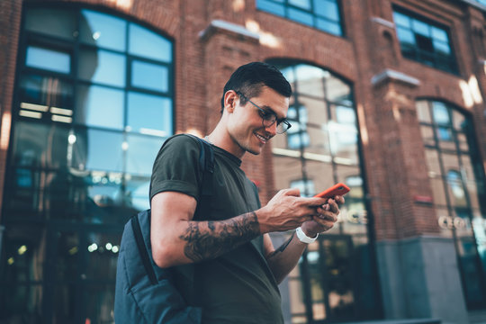 Positive male tourist with backpack for travelling reading received email with funny content information and advices for trip, happy man in eyeglasses checking speed of interent connection on cellular