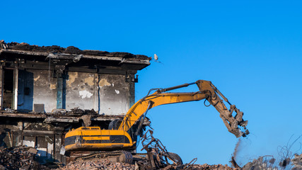 A crawler excavator separates scrap metal from construction debris against the background of a dilapidated building.
