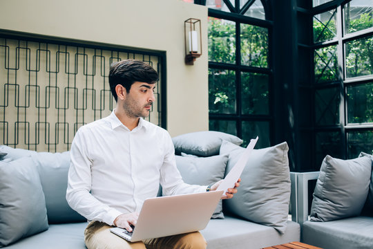 Pondering businessman in elegant wear reading report planning while working remotely on laptop computer, confident spanish amel executive manager holding paper with analytic having freelance job.