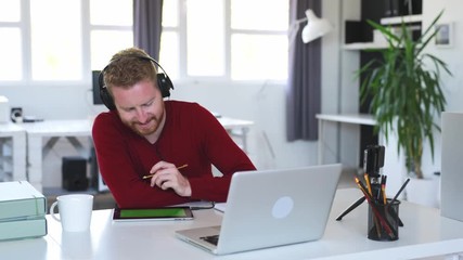 Young handsome innovative caucasian freelancer sitting in his office with headphones on, watching tutorial on tablet and writing in notebook result of his research.