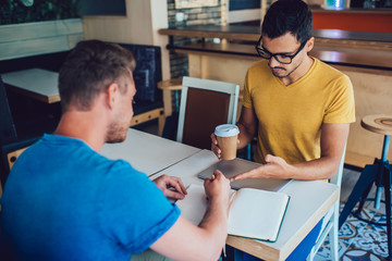 Serious male leader in casual wear and eyewear explaining strategy to employee on coffee break, skilled hipsters guys employees creating plan for cooperation on meeting writing 