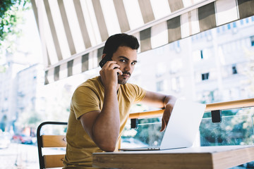 Fototapeta premium Serious caucasian male freelancer making smartphone call while working on cafe terrace remotely using laptop computer, confident man millennial concentrated on mobile phone conversation with operator.