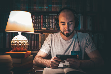 Reading and education. A man in the library is listening to an audiobook, or watching a video on the phone, and there is an open book on the table. In the background is a shelf of books. Close up