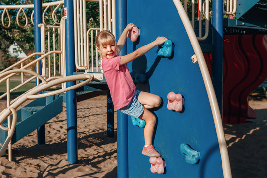 Happy Smiling Little Preschool Girl Climbing Rock Wall At Playground Outside On Summer Day. Happy Childhood Lifestyle Concept. Seasonal Outdoors Activity For Kids.