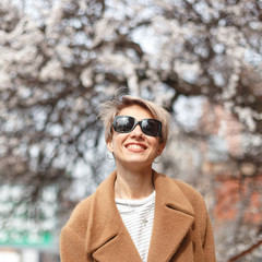 young attractive blonde girl in a coat walks the streets of the spring city with blossoming apple trees. Spring mood, positive emotions, sunny weather.