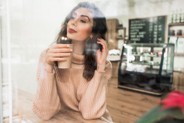 Girl holds a paper cup with coffee. Young beautiful girl is looking through the window.