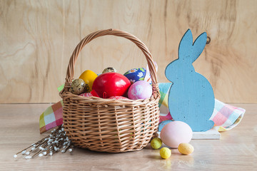 Easter holiday composition. Wicker basket with various easter eggs, bouquet of willow, rabbit and checkered napkin on wooden background