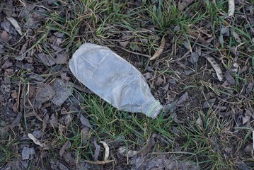trash in nature from one empty white plastic bottle on green grass and gray earth in the park