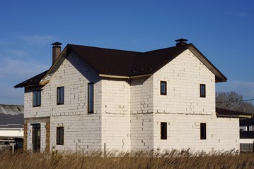 one large private house of white bricks with windows under a brown tiled roof on the street in gray dry grass on a sunny day against a blue sky