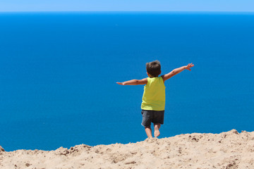 Young male caucasian preschooler jumping from a sandhill into a perfect blue sea.