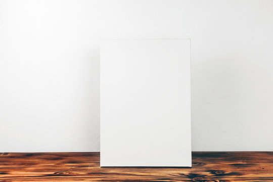 Vertical blank canvas on stretcher standing on table against white wall. Mockup poster frame in modern interior.