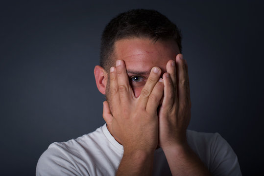 Anxiety - a conceptual image of a beautiful young caucasian man covering his face with his hands and peering out with one eye between her fingers standing indoors. Scared from something or someone.