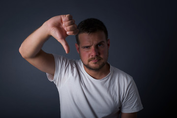 Discontent European man shows disapproval sign, keeps thumb down, expresses dislike, frowns face in discontent, dressed in white shirt, isolated over gray background. Body language concept.