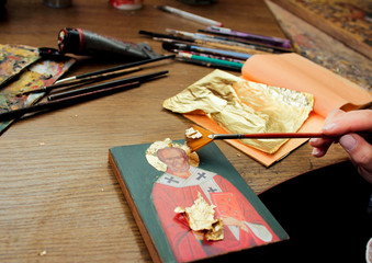 Fototapeta na wymiar Hand with brush of gold leaf. Artist applies gold leaf to the icon. Reproduction of the icon of St. Nicholas. The process of the artist's work. Brushes and paints on the background.