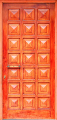 Old orange wooden entrance doors with a bronze handle and symmetrical square panels in Greek style.