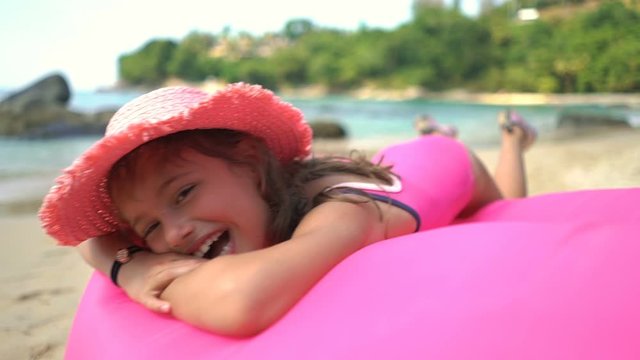 A little happy girl wearing a pink straw hat lying on an inflatable pink sofa on a Sunny beach