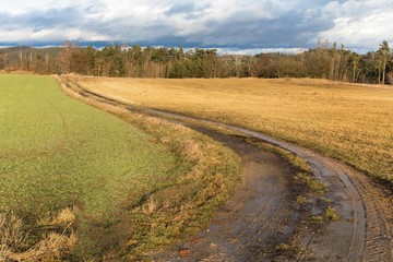 Muddy dirt road in the countryside in the Czech Republic. Dramatic sky. Winter without snow. Road between fields.