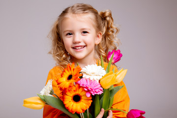 Obraz na płótnie Canvas little blonde girl in an orange dress holds tulips and gerberas in her hands on a white background, a child girl smiles and holds spring flowers in her hands, space for text
