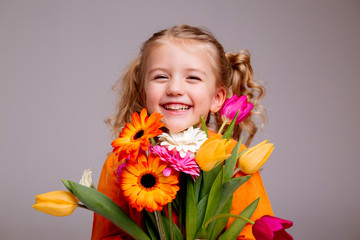 Obraz na płótnie Canvas little blonde girl in an orange dress holds tulips and gerberas in her hands on a white background, a child girl smiles and holds spring flowers in her hands, space for text