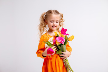 little blonde girl in an orange dress holds tulips and gerberas in her hands on a white background,...