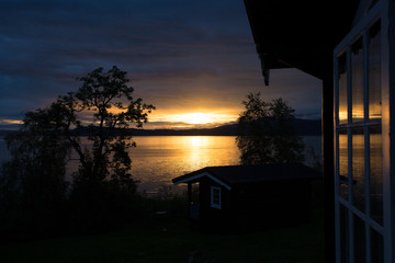Sunrise on 4 a.m. at Trondheim fjord. Sunbeams are reflected in the water and in the window of a small cosy cottage.