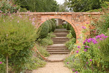 Fototapeta na wymiar A gravel path passes through a circular archway in an old brick wall, which leads on to some old brick steps in an English country garden