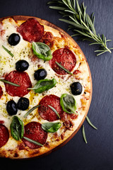 Pizza with salami, mozzarella cheese, black olives, and fresh herbs on black stone background. Top view.