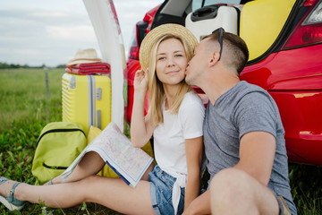 Young married couple sitting in nature at a red car with an open trunk, looking at the map kissing on the cheek